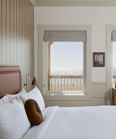 A coastal-themed bedroom features a bed, pillow, framed photo, desk, and ocean view through windows, creating a serene atmosphere.