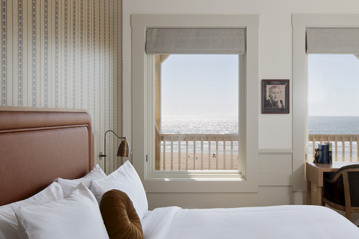 A coastal-themed bedroom features a bed, pillow, framed photo, desk, and ocean view through windows, creating a serene atmosphere.
