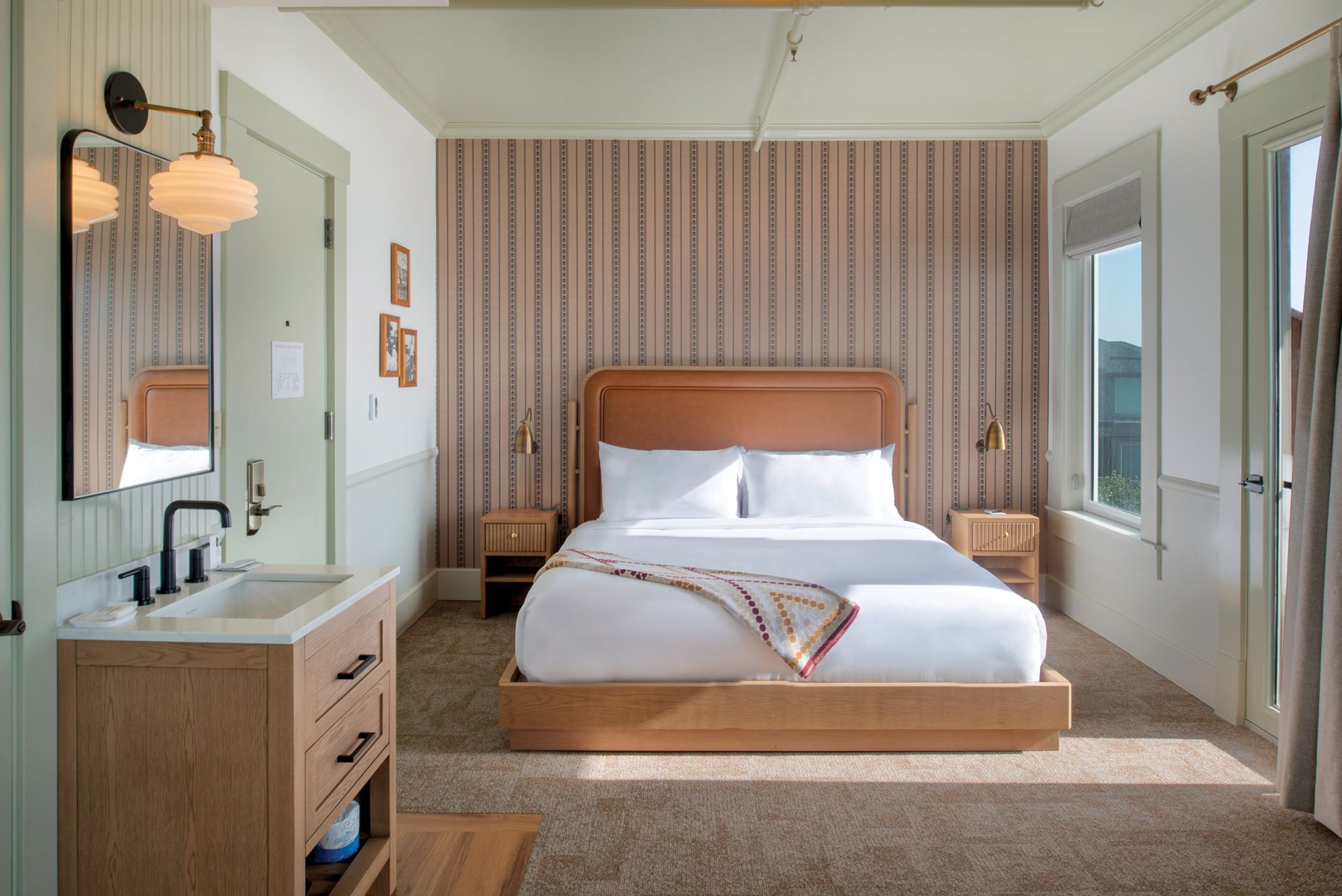 A cozy bedroom with a large bed, nightstands, a wall mirror, and a small sink area. Warm lighting and patterned wallpaper create a charming space.