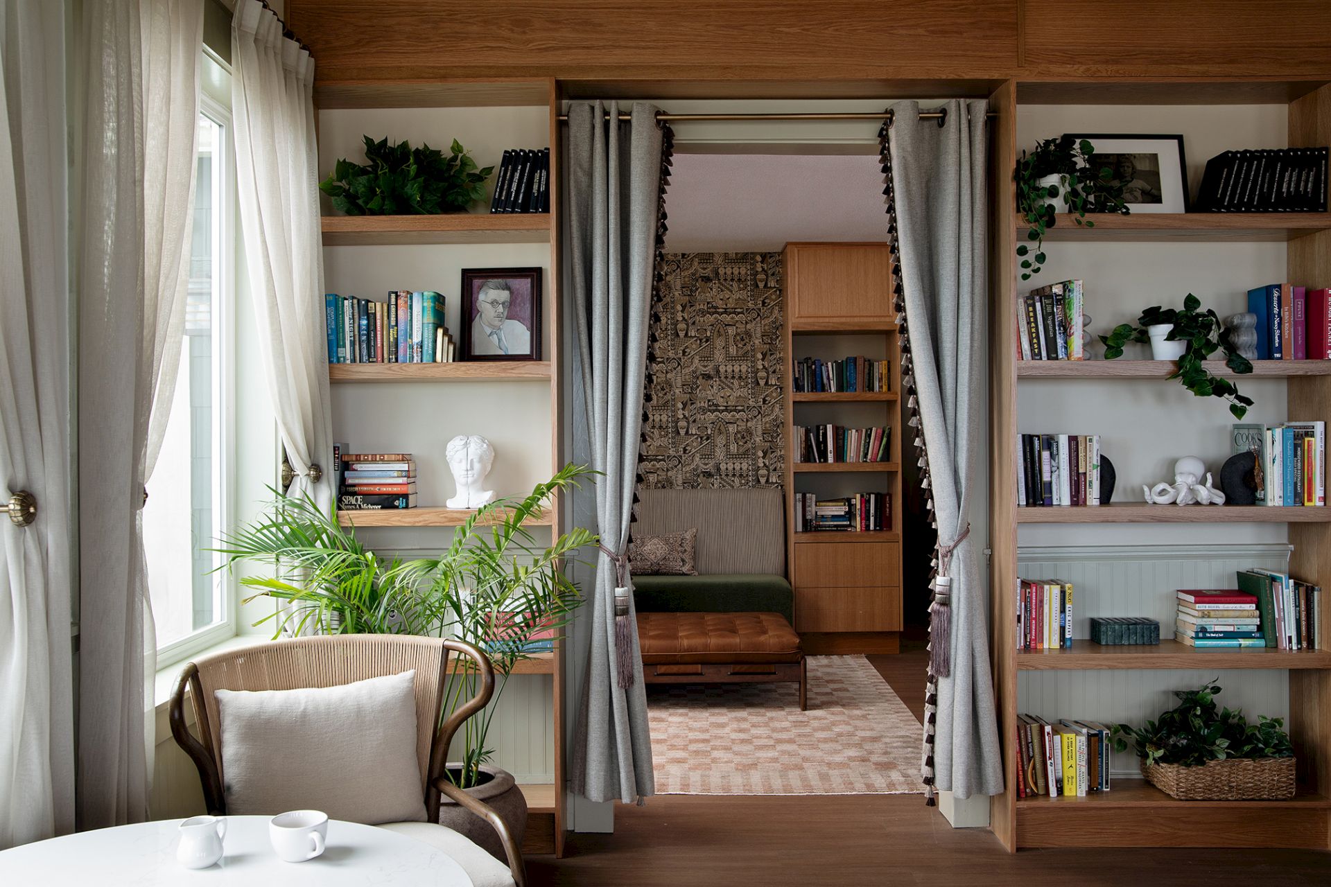 A cozy room nook with built-in bookshelves, grey curtains, and plants, lit by a window and styled with decor.