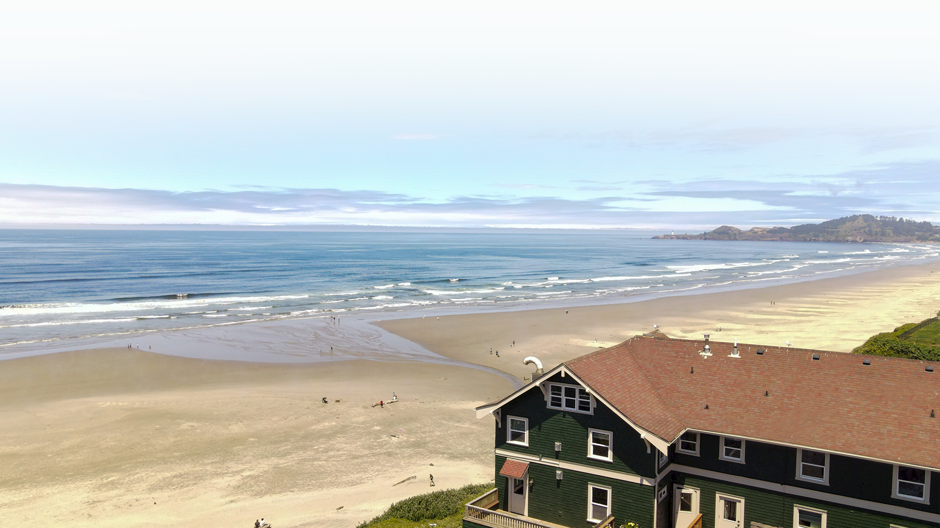 A coastal scene with a sandy beach, ocean waves, and a house with a red roof in the foreground under a partly cloudy sky.