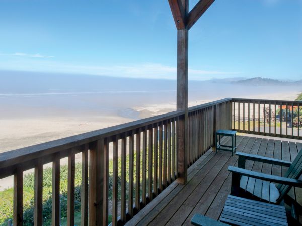 A wooden deck overlooking a sandy beach with two chairs under a clear blue sky, offering a peaceful seaside view.