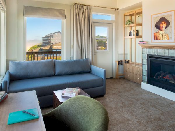 A cozy living room with a blue sofa, fireplace, desk, and artwork on the wall, overlooking an outdoor view through windows and a door.