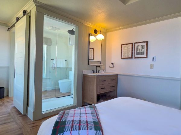 A cozy hotel room with a bed in the foreground, a glass-walled shower, a small desk and mirror, a chair and bench, and soft wall lighting.