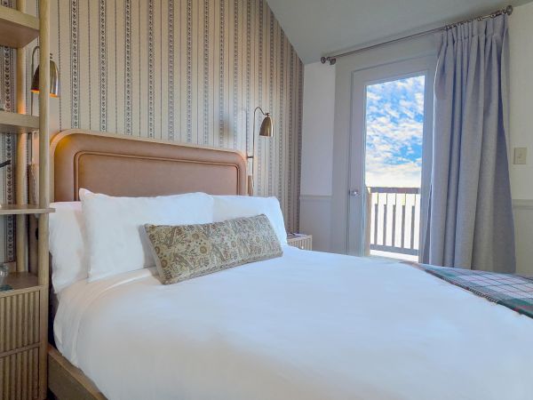 A cozy hotel room with a wooden headboard, white bedding, patterned throw pillow, built-in bookshelf on the left, and a balcony door letting in light.
