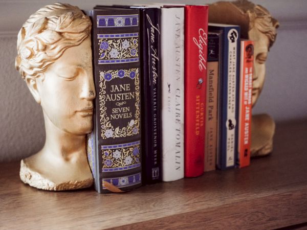 A carved bust with a row of books&mdash;classic novels and literature spines&mdash;lining a wooden shelf, blending art with reading material.
