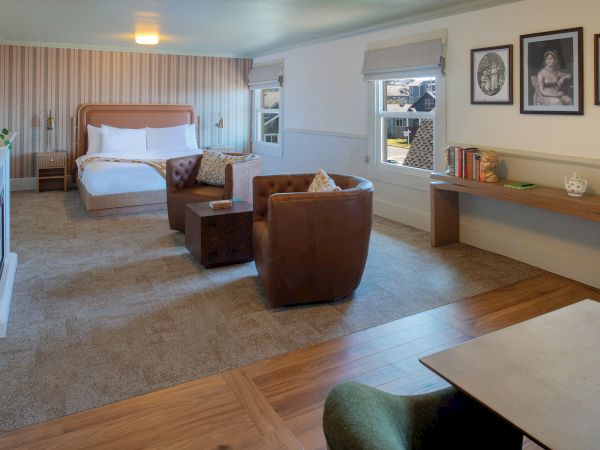 A spacious hotel suite with a king bed, two brown chairs, a sofa, desk area, and wall art; warm lighting and wood flooring.
