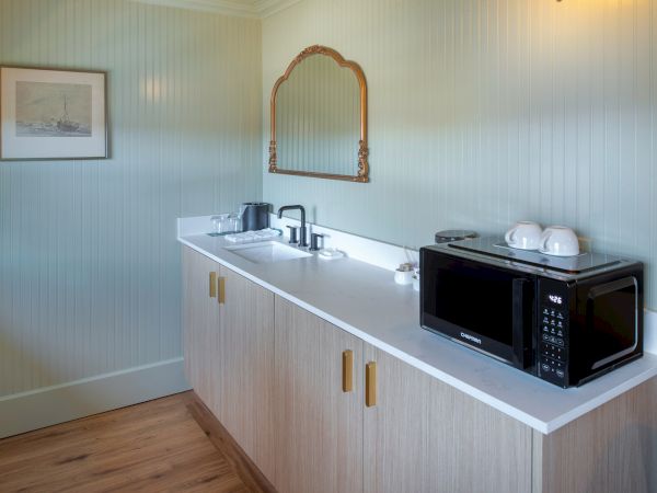 A compact kitchenette with a long white counter, light wood cabinets, a microwave, kettle, cups, a sink, a framed mirror, and pale blue walls. Ending with a period.