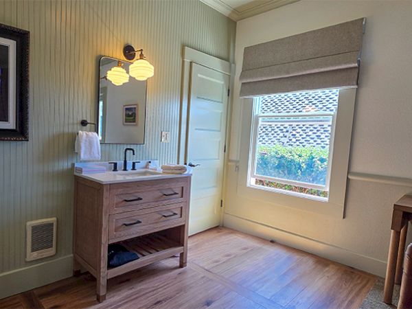 A bathroom with a wooden vanity, a white sink, wall-mounted light, framed Purple Rain poster, beamed mint-green walls, a window with a shade, and a small chair in the corner.
