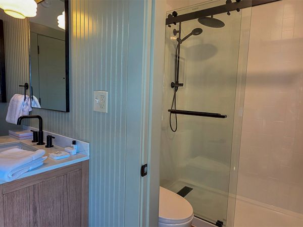 A small bathroom with a wooden vanity, white sink, wall mirror, towel rack, and a glass-enclosed shower with a handheld wand, all in a light blue-green paneled room.