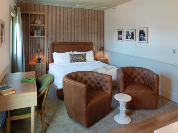 A cozy hotel room with a king bed, brown tufted armchairs, a small white side table, a desk and chair, warm wood tones, and framed photos on the wall.
