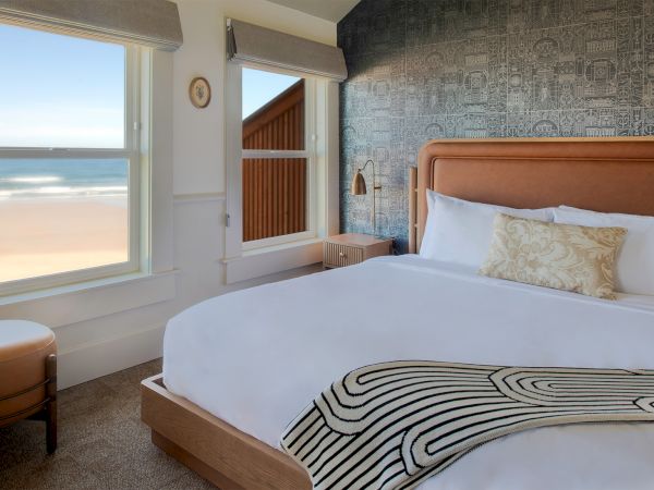 A cozy bedroom features a large bed, decorative throw, patterned wall, and windows with a beach view.