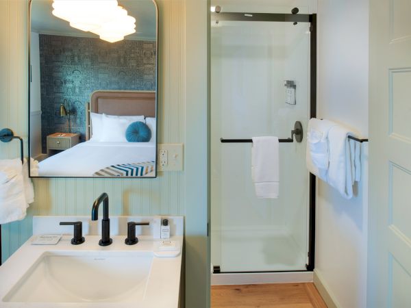 A bathroom with a sink, mirror, soap, and black fixtures. Next to it, a glass shower with a towel hangs on the door handle.