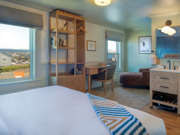 A cozy room with a bed, desk, and chair, featuring a window view, open shelving, an armchair, and a small vanity area.