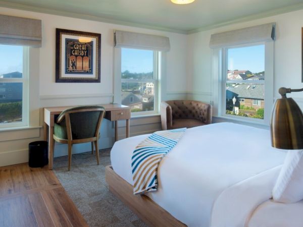A cozy bedroom with a large bed, striped blanket, desk, chair, and window views. The decor includes artwork and light-colored walls.