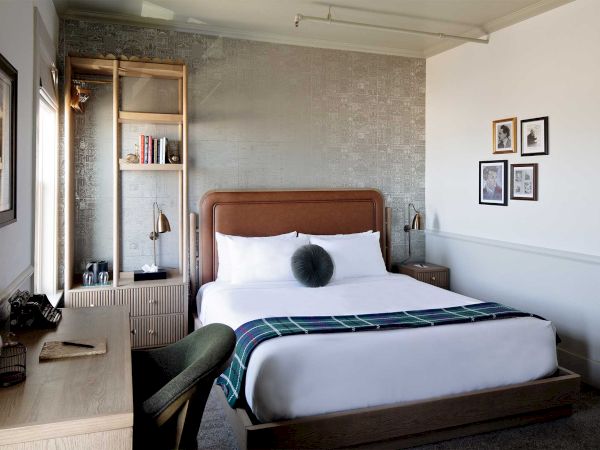 A cozy bedroom with a neatly made bed, decorative pillows, a blanket, desk, chair, lamp, shelves, and framed pictures on the wall.