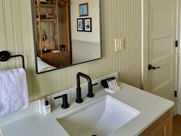 A bathroom scene with a white sink, black faucet, wall mirror, and two hanging globe lights; a cabinet and framed art reflect in the mirror, with soft beige paneled walls.