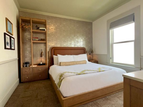 A cozy bedroom with a wooden bed, white linens, a metallic accent wall, a built-in bookshelf, and a large window letting in natural light.