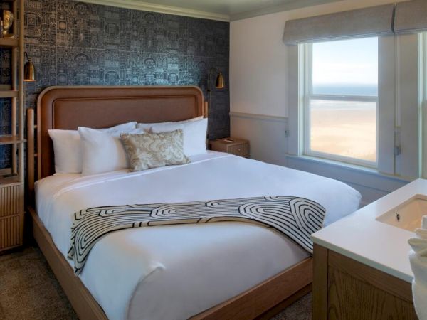 A cozy bedroom with a large bed, patterned throw, and ocean view through the window. Shelves with decor are on the left wall.