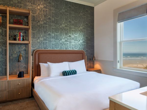A cozy bedroom with a double bed, decorative wallpaper, and a window view of the ocean, complemented by a wooden shelf and warm lighting.