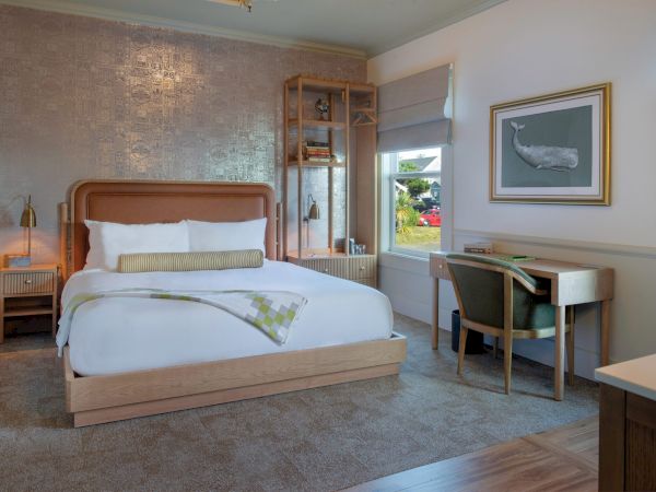 A well-lit hotel room features a bed, desk with a chair, decorative wall art, and a window showing an outside view.