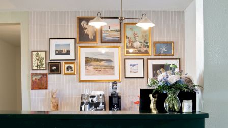 A dark green bar with a brass foot rail sits in front of a wall gallery of framed art; a couple of hanging lights illuminate the colorful collage above.