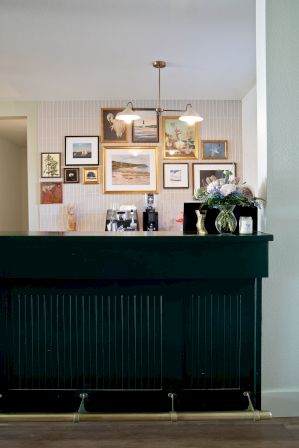 A dark green bar with a brass foot rail sits in front of a wall gallery of framed art; a couple of hanging lights illuminate the colorful collage above.