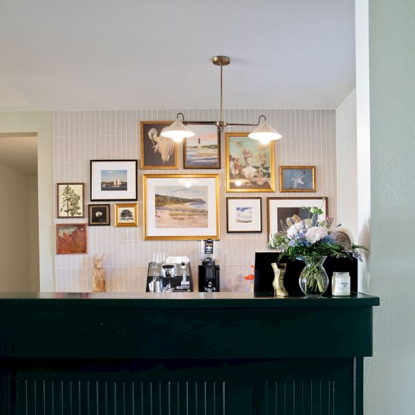 A dark green bar with a brass foot rail sits in front of a wall gallery of framed art; a couple of hanging lights illuminate the colorful collage above.