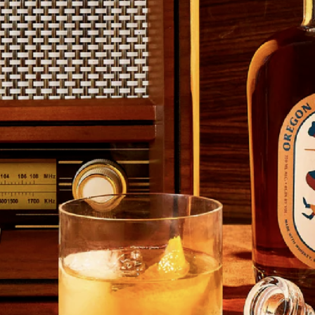 A glass of whiskey with ice and a lemon twist sits on a wooden surface, next to a bottle and a vintage radio.