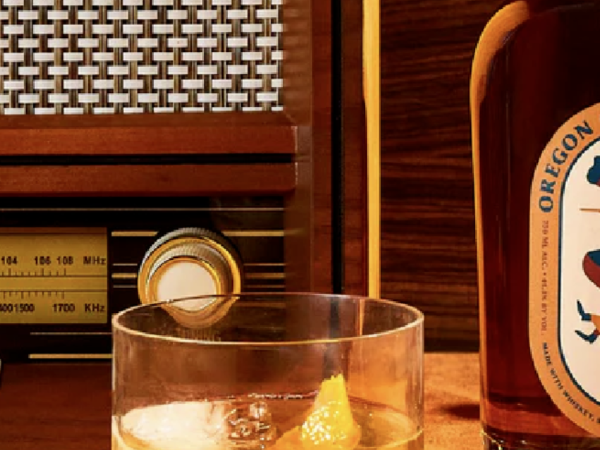 A glass of whiskey with ice and a lemon twist sits on a wooden surface, next to a bottle and a vintage radio.