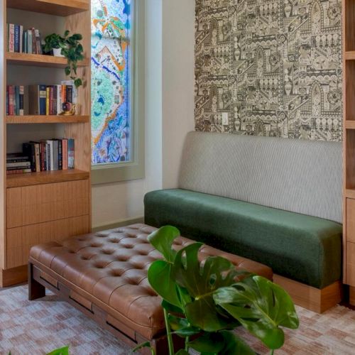 A cozy room with bookshelves, a bench, a plant, and a piano. A decorative wall panel and a colorful window add charm to the space.