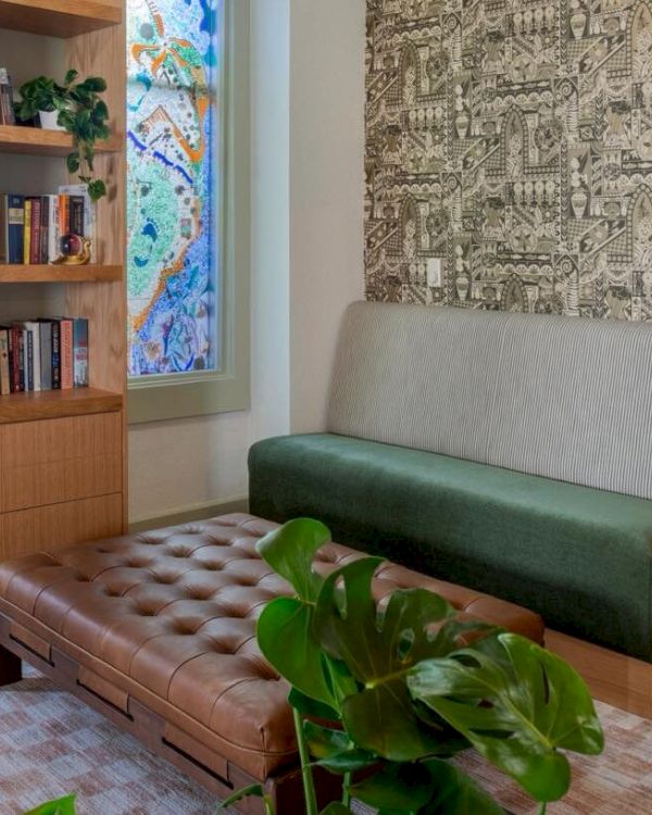 A cozy room with bookshelves, a bench, a plant, and a piano. A decorative wall panel and a colorful window add charm to the space.