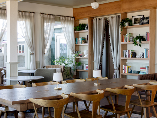 A cozy room with a large wooden table, chairs, bookshelves, and soft lighting, featuring plants and curtains for a warm ambiance.