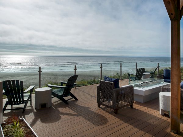 A patio with chairs, a table, and a fire pit overlooks a beach and ocean under a partly cloudy sky.