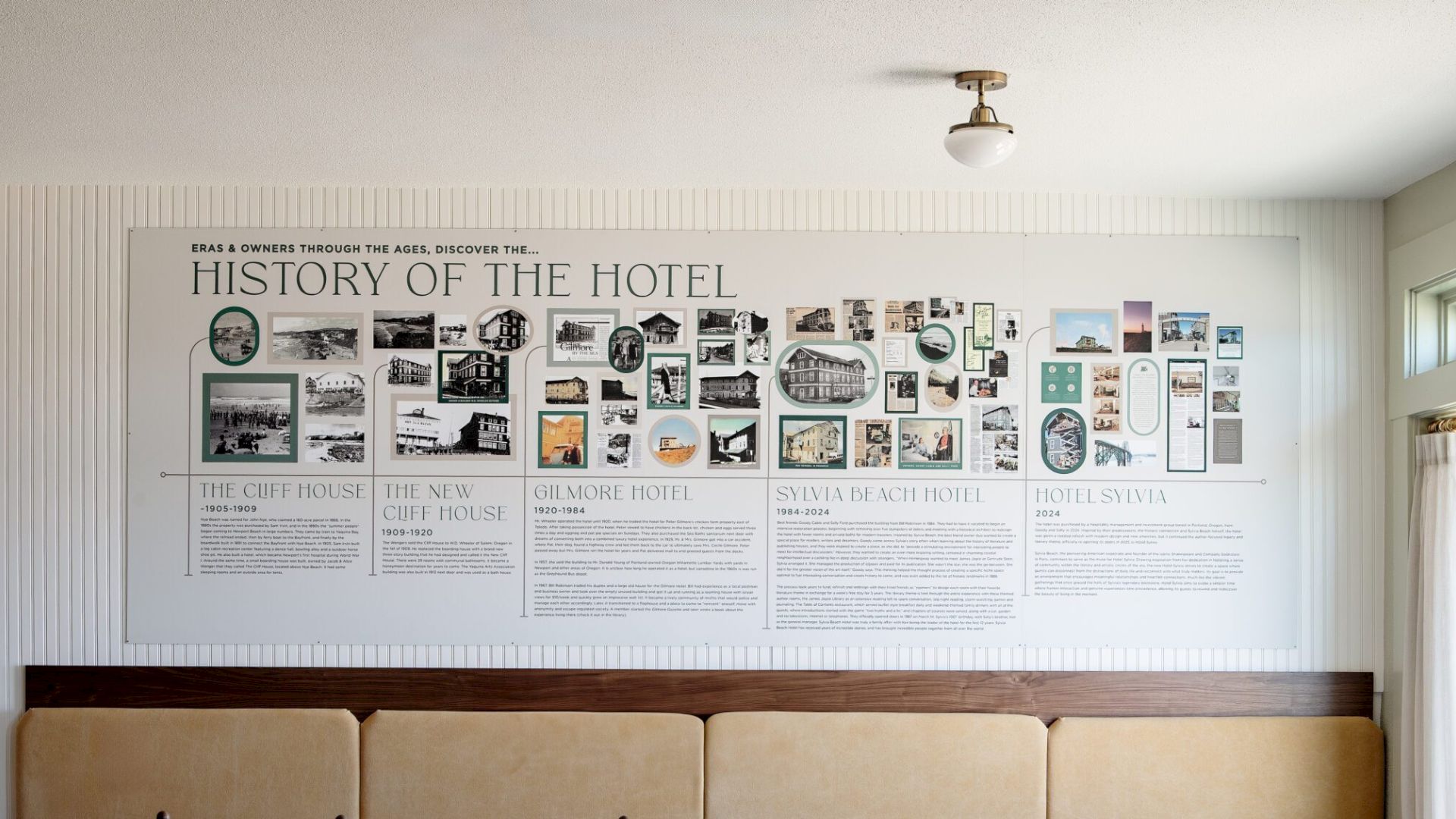 A retro hotel lobby wall with a long timeline poster titled &ldquo;History of the Hotel,&rdquo; small photos and text panels above a tan cushioned bench.
