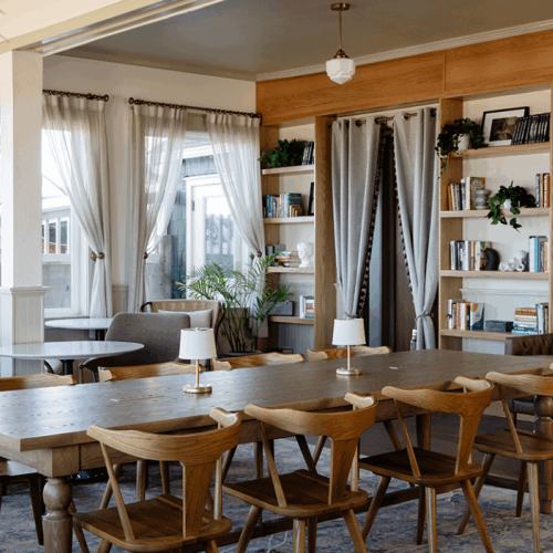 A cozy dining room with wooden chairs, a large table, bookshelves, and soft lighting. Curtains frame the windows for a homey feel.