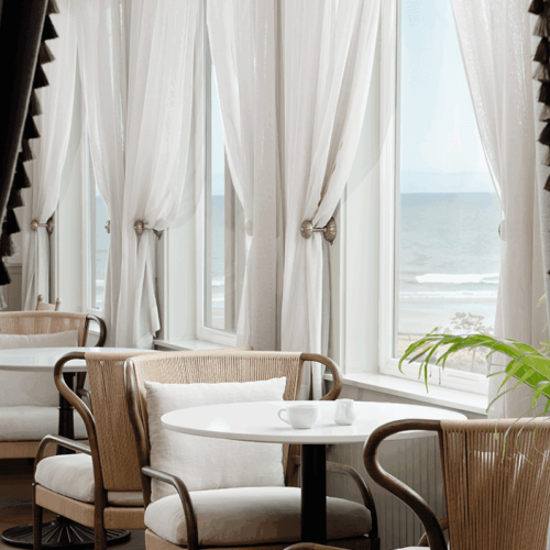 A cozy caf&eacute; with white curtains, a round table, and chairs overlooks the ocean through large windows, creating a serene atmosphere.