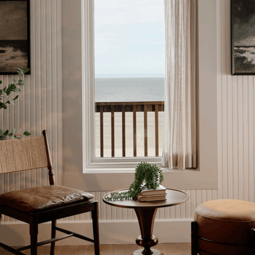 A cozy room with two chairs, a small table with books and a plant, and a window with an ocean view and framed art on walls.