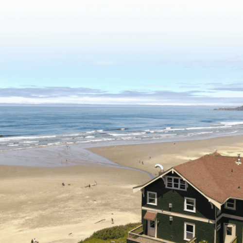 A coastal scene with a sandy beach, a green building with a red roof, and a distant headland under a clear blue sky.