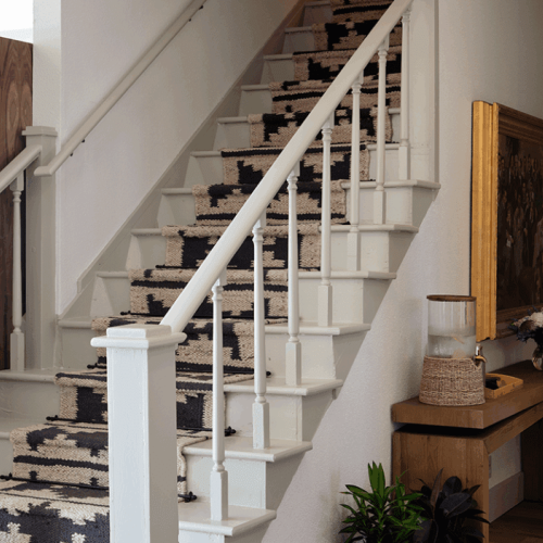 A staircase with a patterned runner, white railing, and a nearby console table with decor and a painting.