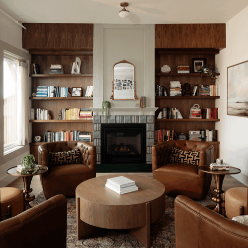 A cozy living room features leather chairs, a circular table, a fireplace, and bookshelves filled with books and decor.