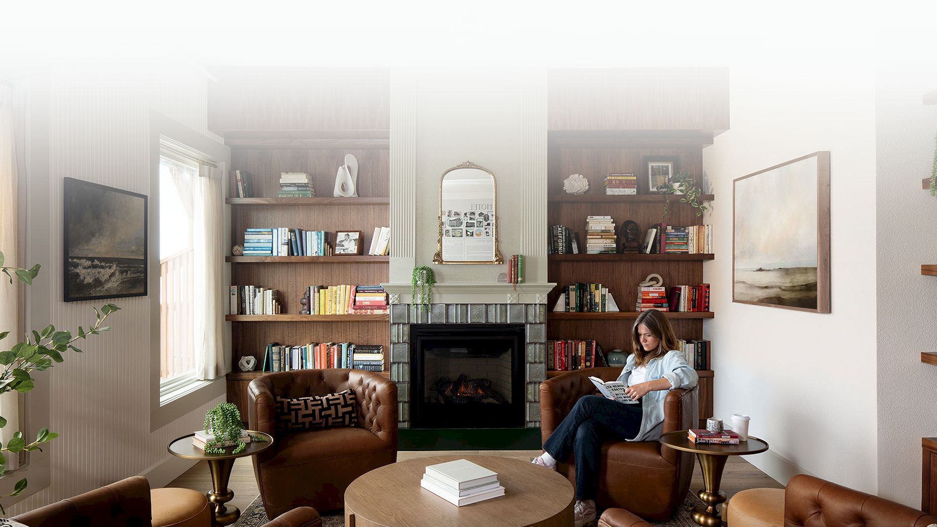 A cozy living room with bookshelves, a fireplace, and a person reading on a chair. Shelves are adorned with books and decor items.