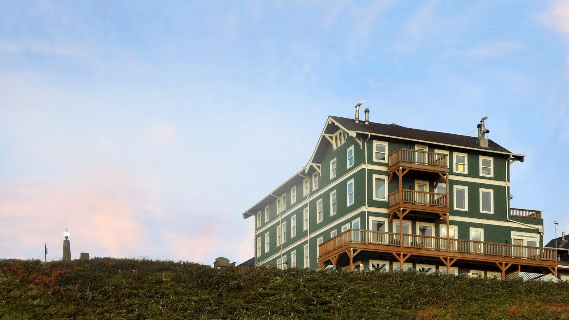 A large green wooden house on a hill with balconies overlooks a grassy slope under a blue sky, a lone lighthouse visible in the distance.