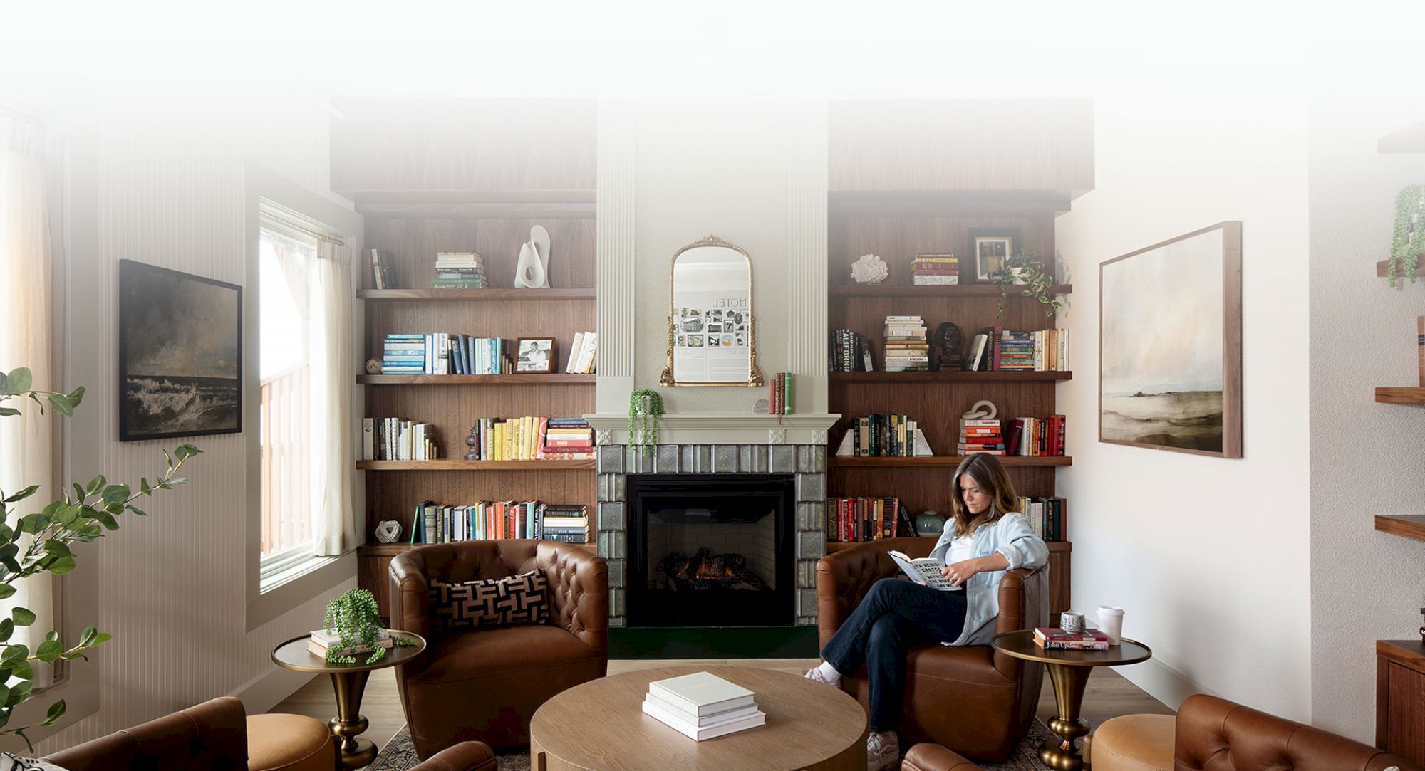 A cozy living room with bookshelves, a fireplace, and a person reading on a chair. Shelves are adorned with books and decor items.