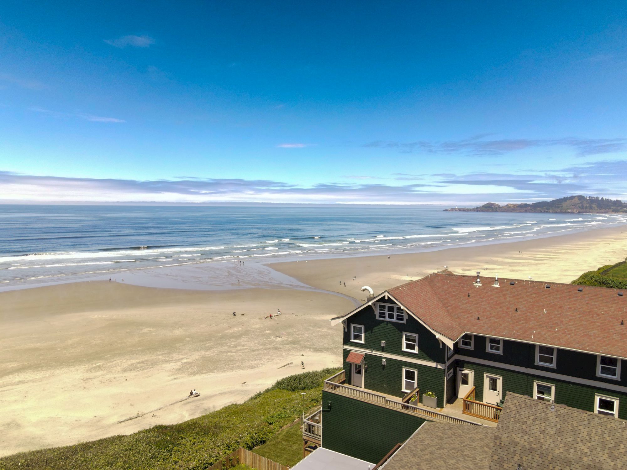 A beach view shows a house with a red roof, sandy shore, ocean waves, and distant hills under a clear blue sky.