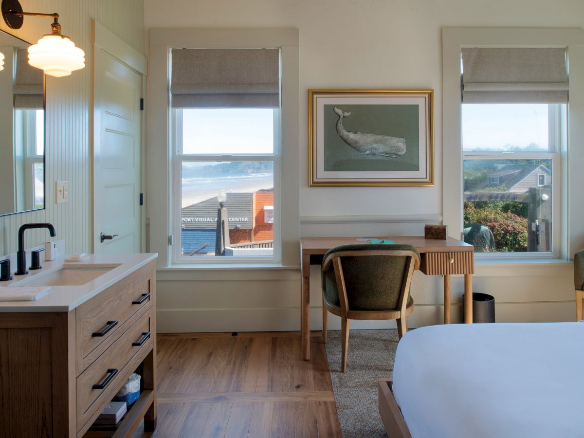 A bright hotel-style room with a wooden vanity, sink, and fixtures on the left, a desk and chair by two windows, a whale painting, and a bed partially visible on the right, with ocean views through the windows.