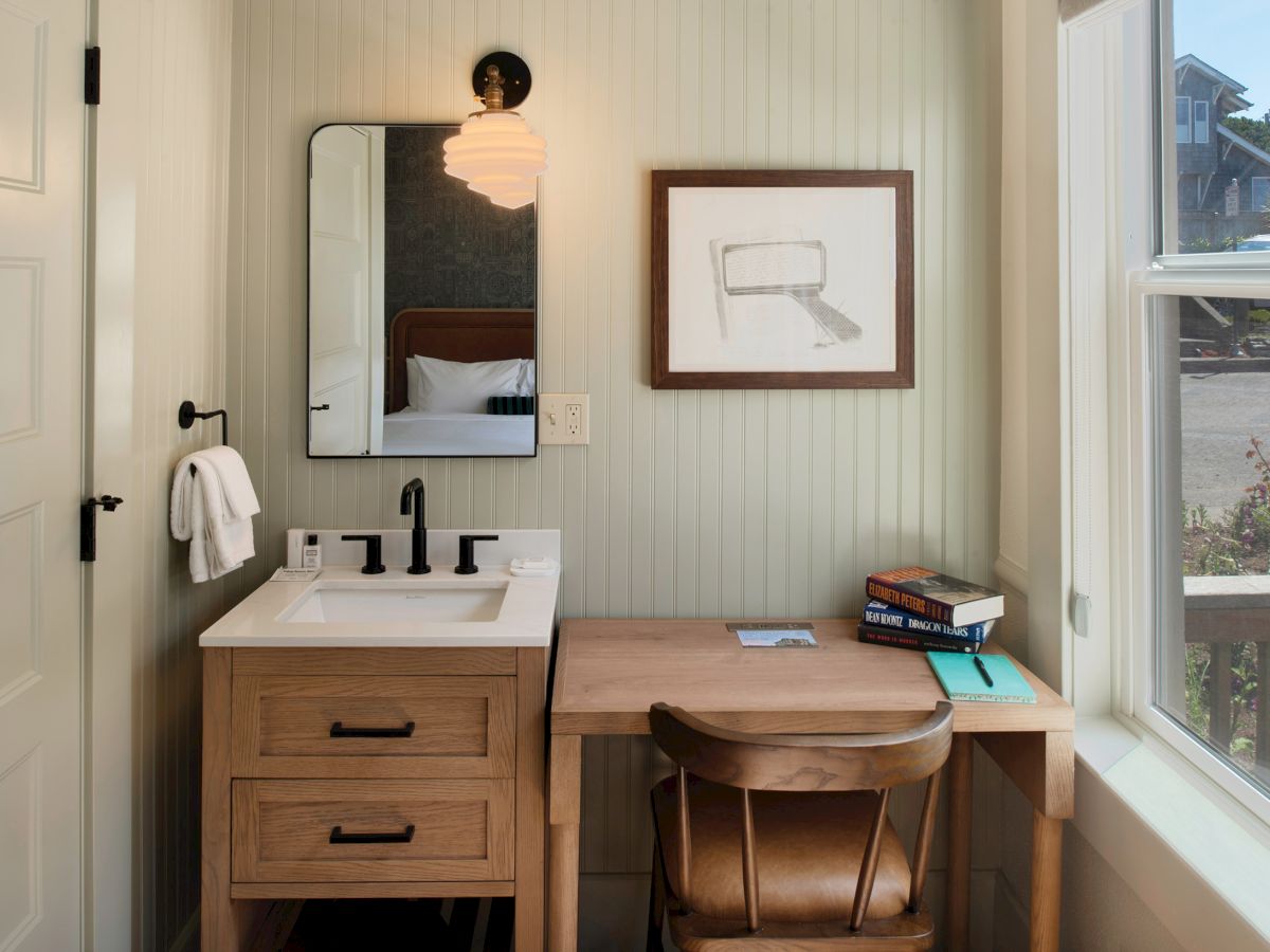 A compact bathroom/desk combo with a small vanity, mirror, wall art, wooden desk and chair, books, and a large window letting in light. Top it at 140 characters, always ending the sentence.