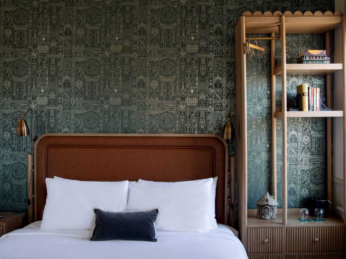 A stylish hotel bedroom with a wooden headboard, white bed linens, a dark throw pillow, and a tall wooden open shelf unit by a green patterned wallpapered wall.