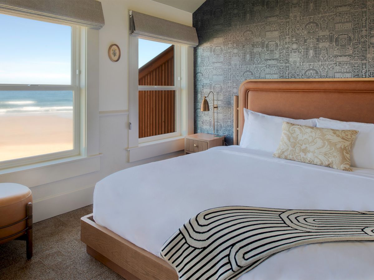 A bright hotel room with a large bed, white linens, patterned throw, sea view through two windows, and a cozy seating nook by the window.