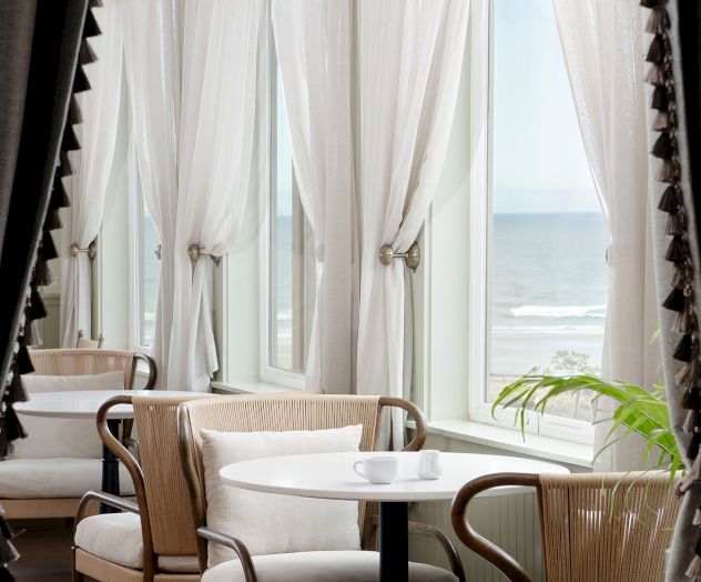 Cozy seaside cafe with white curtains, round tables, woven chairs, and sunlight streaming in by a large window overlooking the water.